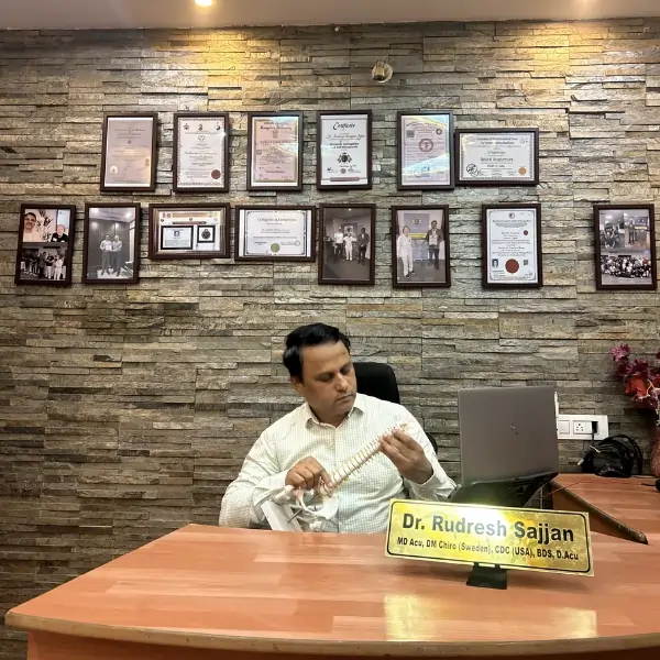 Dr. Rudresh, Europe & US Certified Chiropractor, sitting in his AcuSTAR Wellness consultation room displaying professional diplomas and holding a spine model for patient education in Bangalore